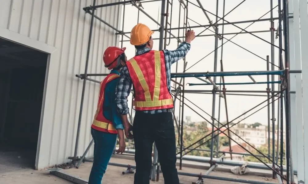 Two workers in safety helmets and reflective vests assembling scaffolding beside a building