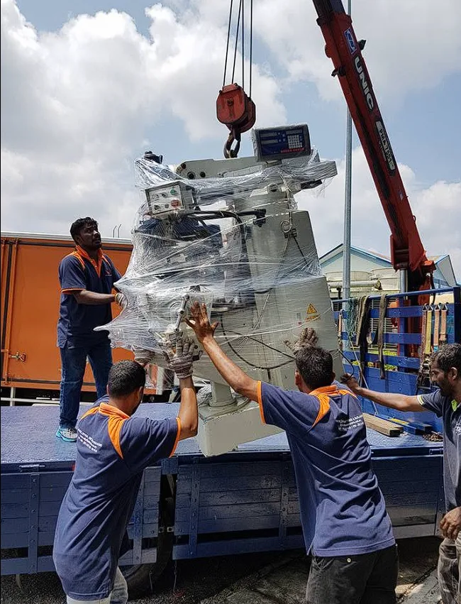 Crane lowering a wrapped milling machine onto a lorry while crew guide it, machinery mover Klang